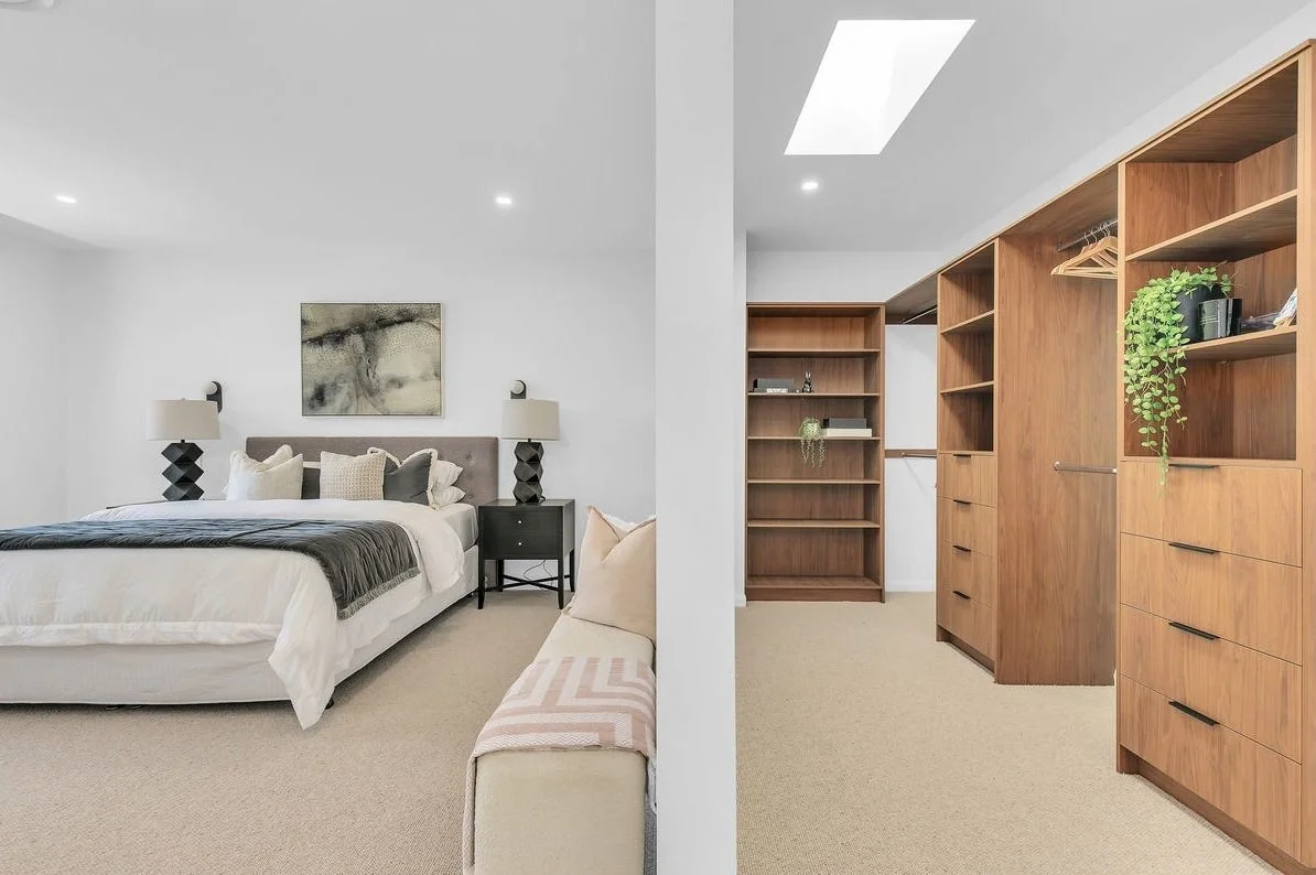 A bedroom with a bed, nightstands, lamps, and wall art on the left, and a walk-in closet with wooden shelves and drawers on the right.