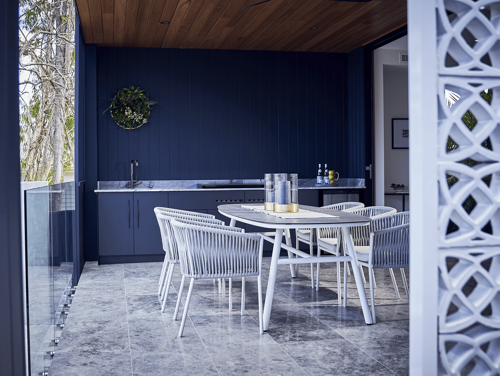 Modern outdoor dining area with a dining table, six chairs, and a built-in outdoor kitchen with a dark blue wall, marble countertops, and a decorative wreath on the wall.