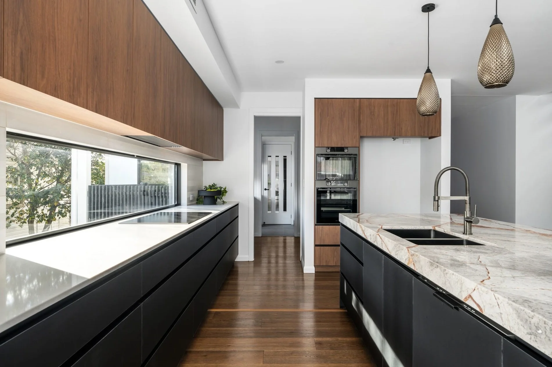 Modern kitchen with wooden cabinetry, a marble island with a built-in sink, black lower cabinets, a long horizontal window, and two pendant lights hanging from the ceiling.