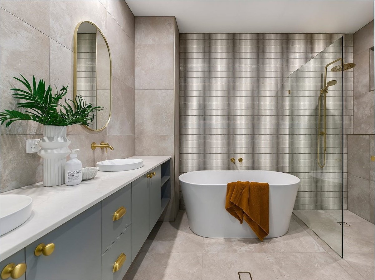 Modern bathroom with double sinks, a large mirror, a potted plant, a freestanding bathtub with an orange towel, and a walk-in shower with gold fixtures and a glass divider.