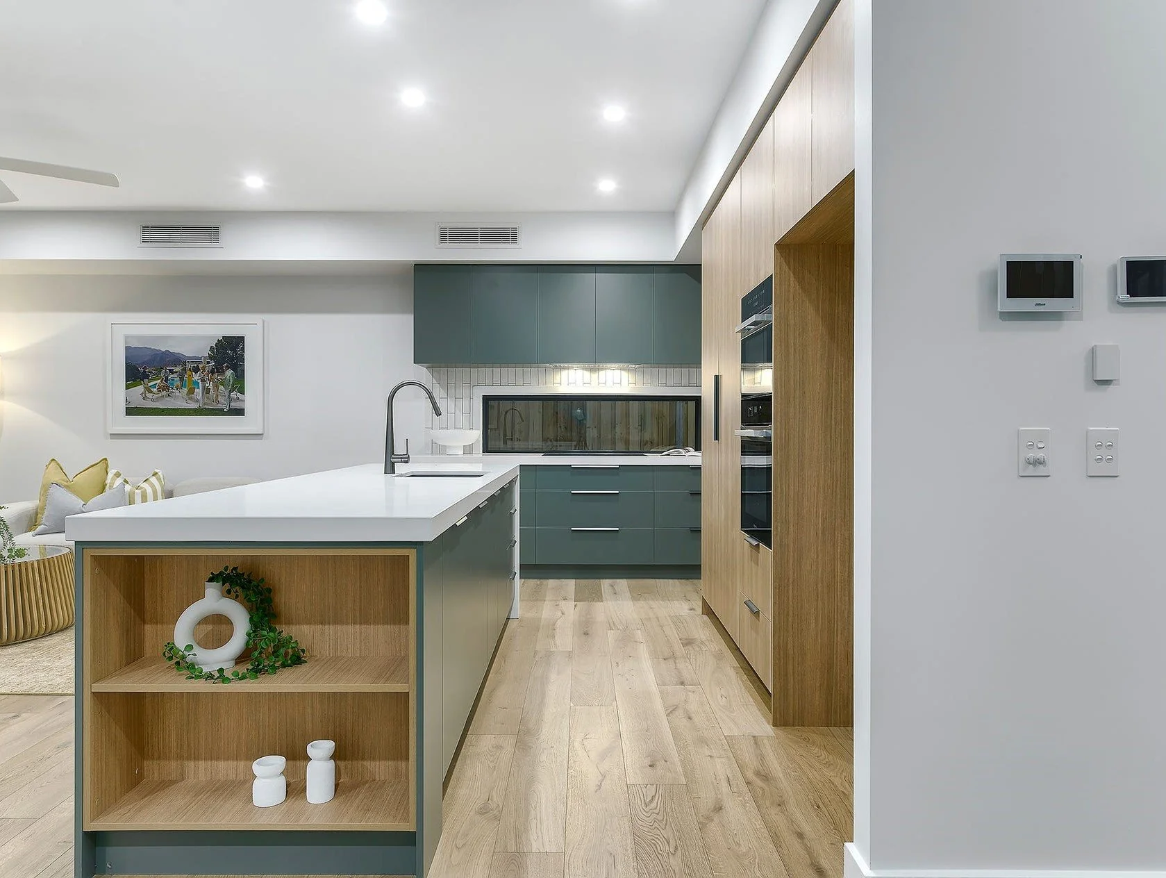 A modern kitchen with a white island, green cabinets, and wooden accents. There's a window behind the countertop, a sink, and built-in appliances in the cabinetry. The room has wooden flooring and a cozy, stylish decor.