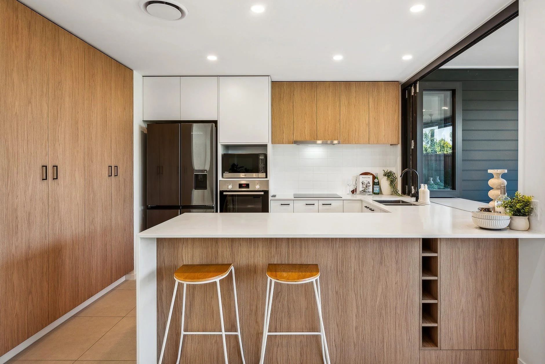Modern kitchen with wood cabinets, white countertops, stainless steel appliances, and a window with a view outside.