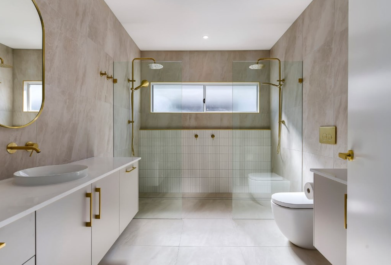 Modern bathroom with beige tiles, gold fixtures, a white vanity with a vessel sink, mirror, and glass-enclosed shower area with two showerheads. There is a window and a white toilet with a toilet paper holder.