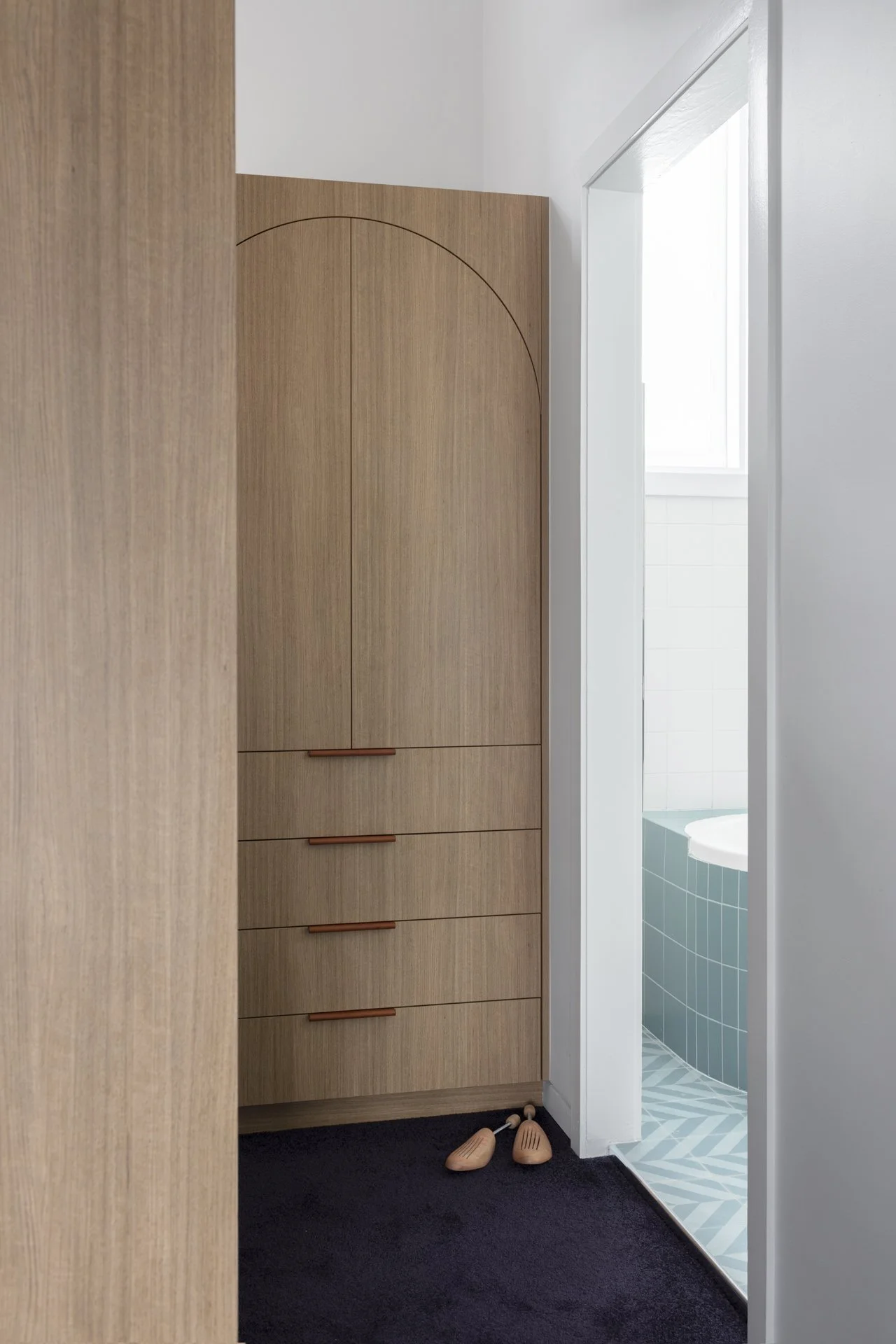 Narrow view of a wooden wardrobe with rounded top doors and horizontal handles, next to a white wall and bathroom with blue tiles and window, on dark carpeted floor.