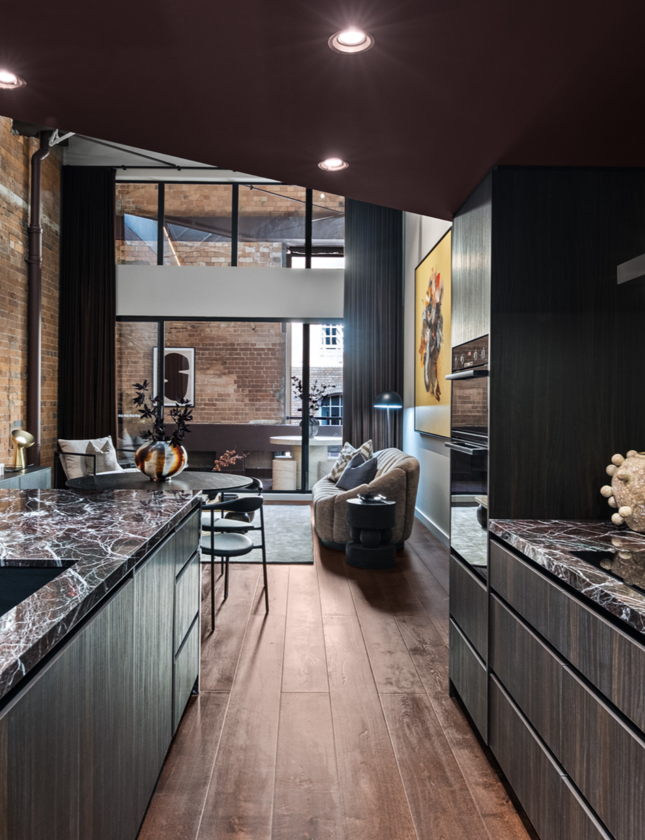 Modern open-concept living area with dark wood cabinets, a marble-topped kitchen island, a sitting area with a beige sofa, a large yellow abstract painting, and floor-to-ceiling windows with brick walls in the background.