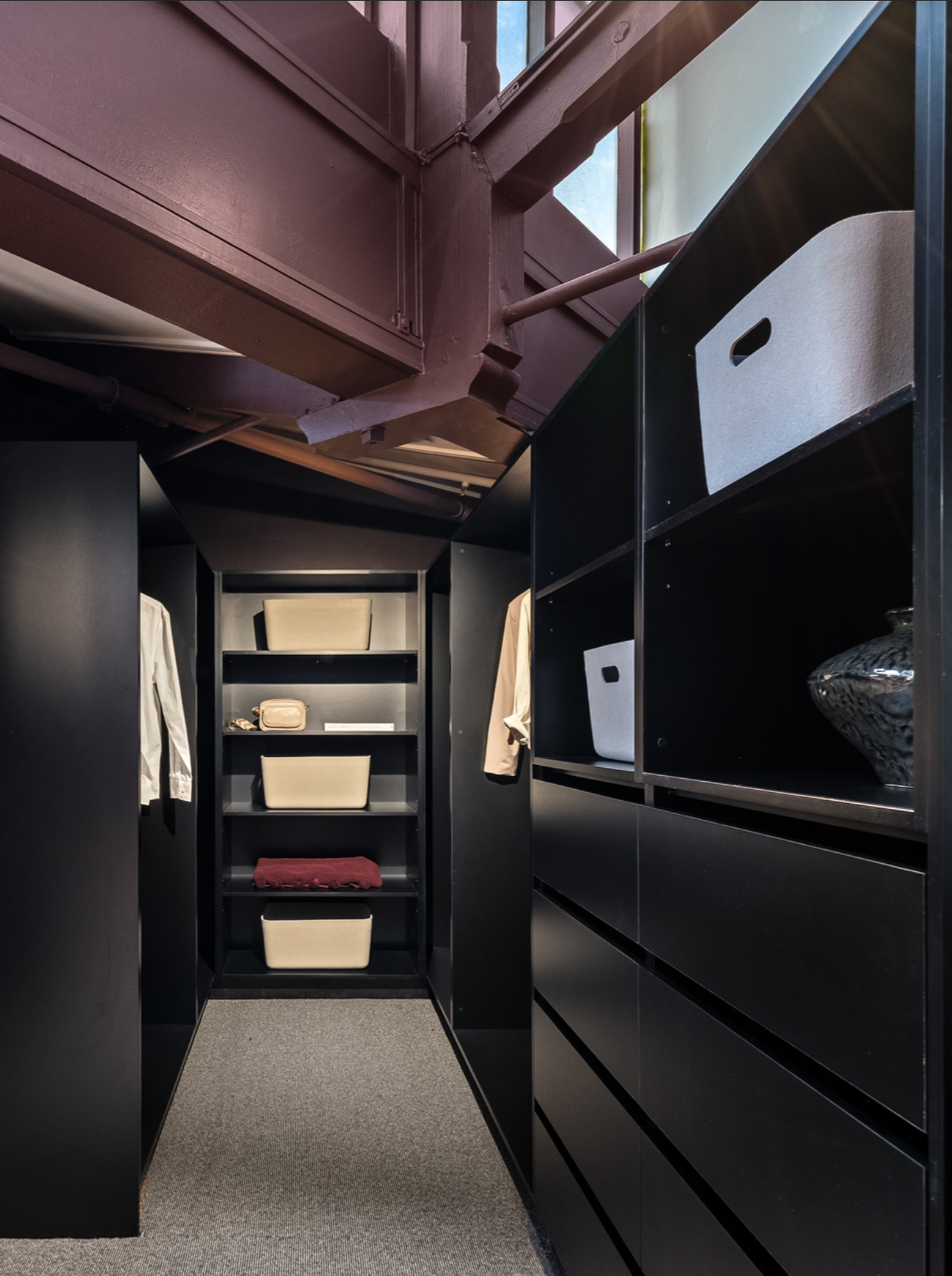 A modern walk-in closet with black shelving units, beige storage bins, and clothing hanging on the sides; there is a small window at the top and exposed wooden beams overhead.