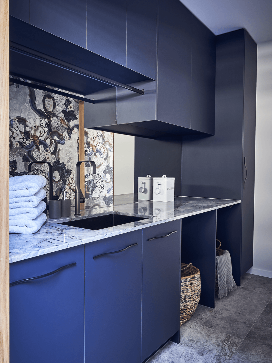 Dark blue kitchen with marble countertop, black sink, black faucet, and cabinet doors. Towels stacked by the sink, decorative painting or wallpaper behind, and baskets stored underneath.