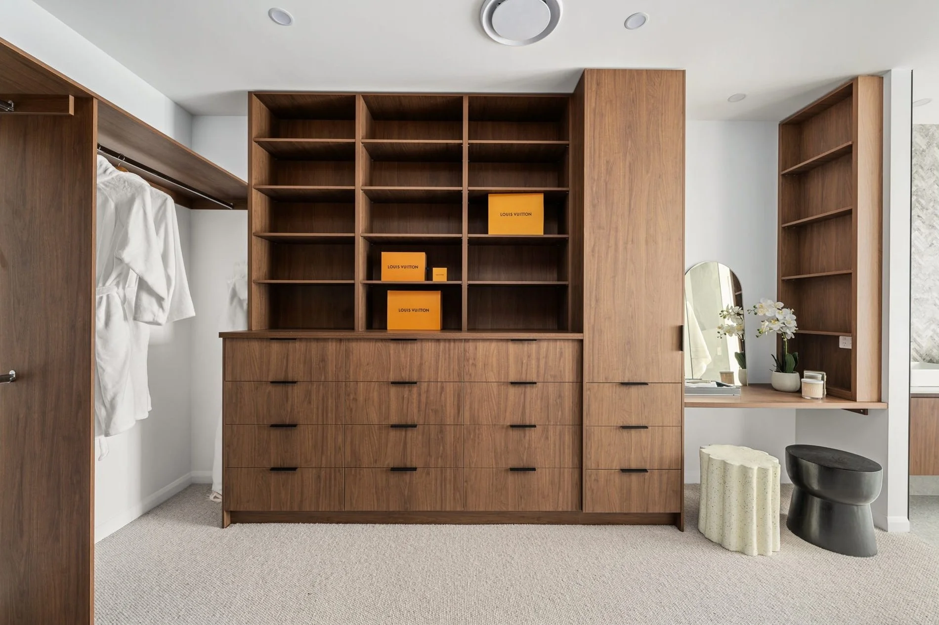 A modern bedroom closet with wooden sliding doors, open shelves, and a dresser with multiple drawers. On the left, a robe hangs on the closet rod. To the right, a vanity area features a mirror, a white orchid, and decor items. There are geometric sto