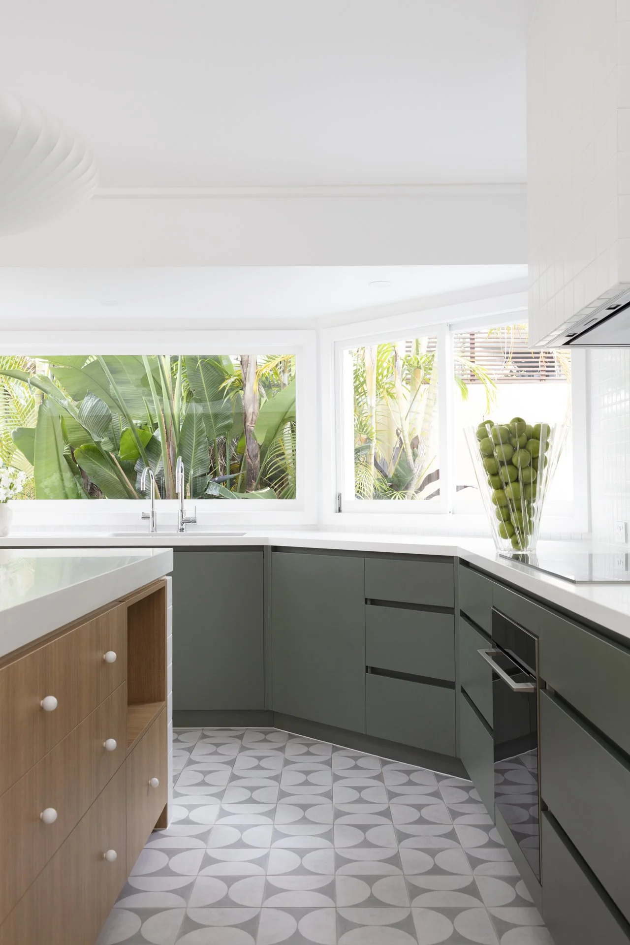 Modern kitchen with white countertops, green cabinets, a large glass vase filled with green apples, and tropical plants visible outside through large windows.
