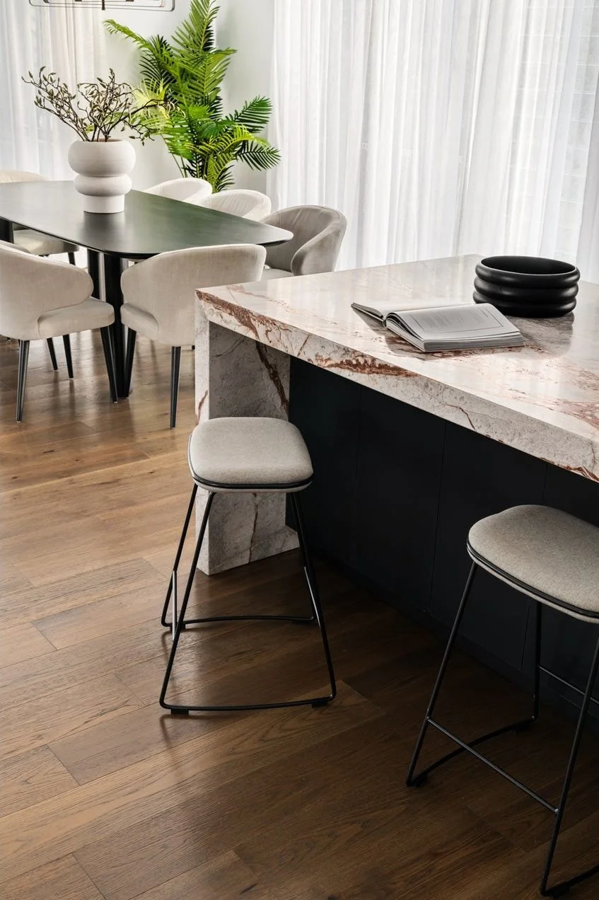 Modern dining and kitchen area with a marble countertop island, beige upholstered stools, a dining table with chairs, potted plants, and light curtains in the background.