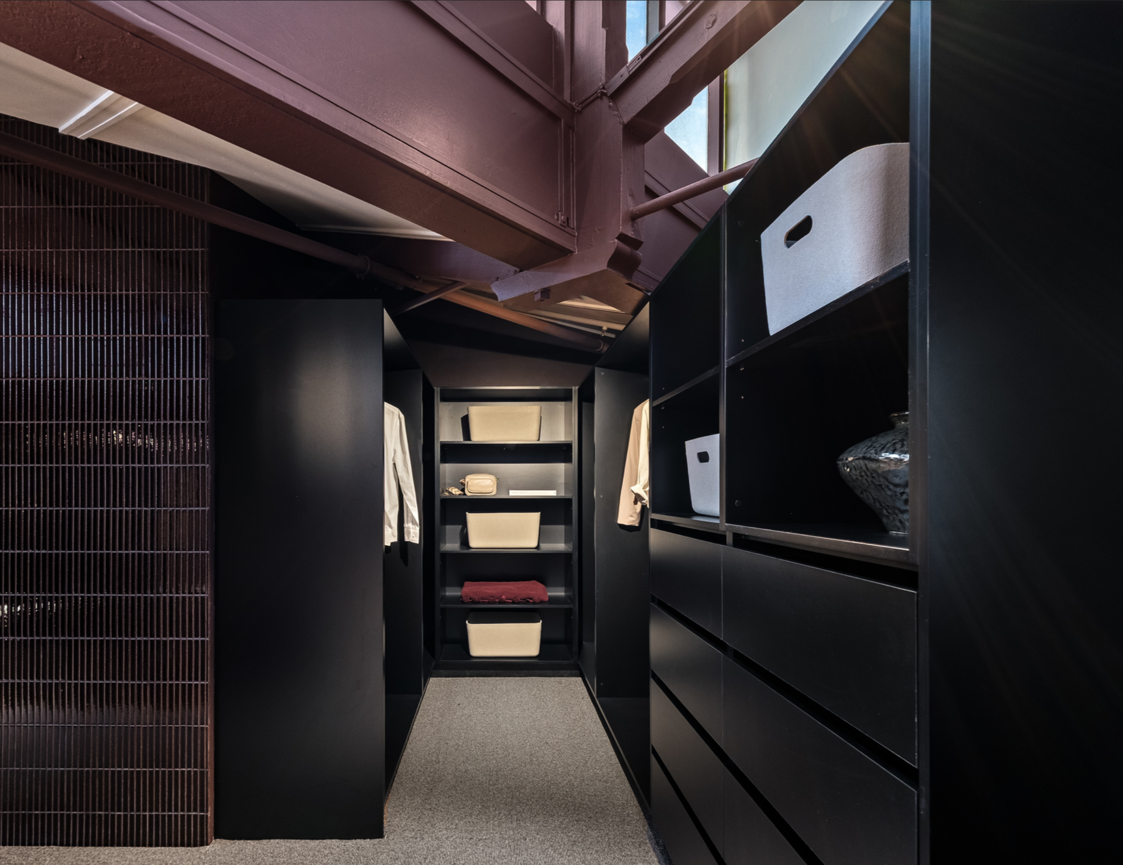 A walk-in closet with black shelves and storage bins, cream and beige clothing hanging, and a window letting in natural light.