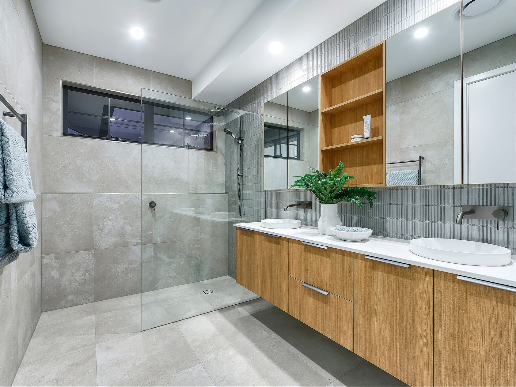 Modern bathroom with double sinks, wooden cabinet, large mirror, potted plant, glass shower enclosure, tiled walls and floors, window, and recessed ceiling lights.