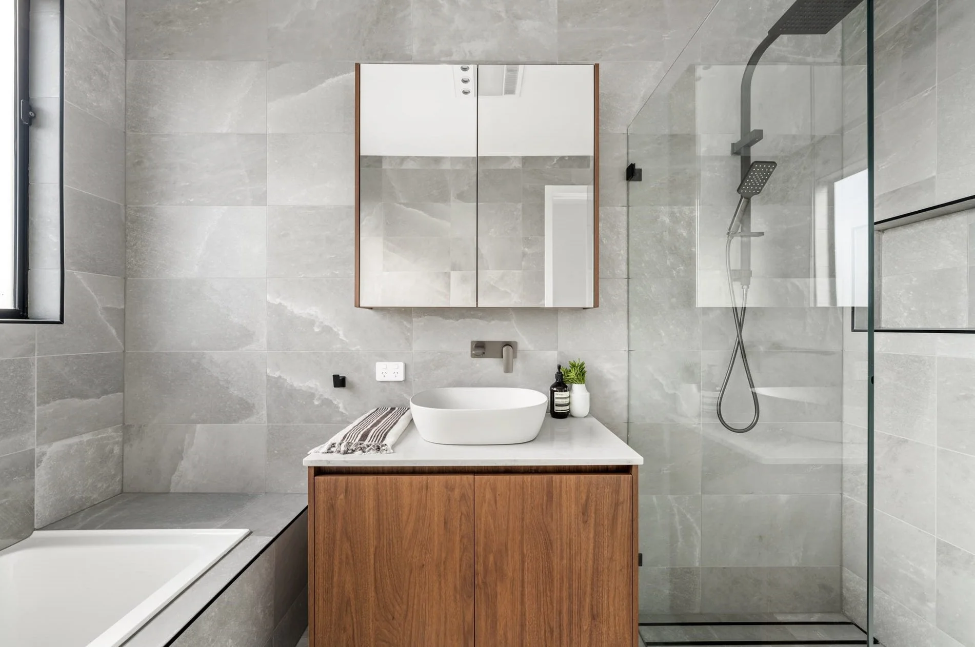 Modern bathroom with gray tiled walls, wooden vanity with a white countertop, vessel sink, and black faucet. There is a mirrored cabinet above the sink, a shower area with a black shower head and glass partition, and a small window on the left.