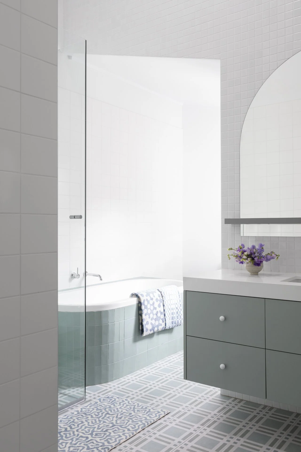 A clean and modern bathroom with a trendy light green vanity, a white countertop, a wall mirror, a vase of purple flowers, a bathtub with two patterned towels, and a geometric-patterned floor.