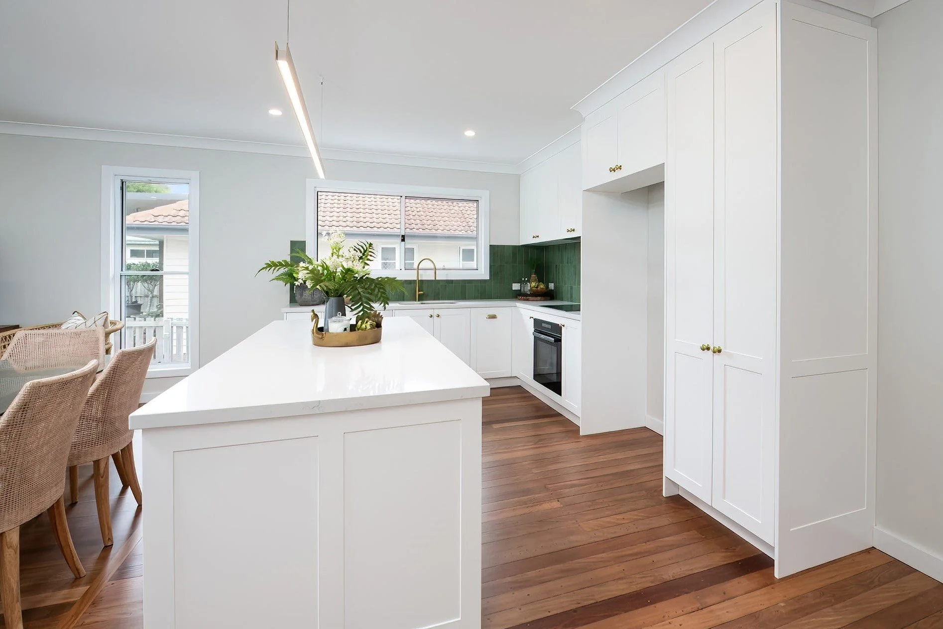 Modern kitchen with white cabinets, a white island with plant decor, wooden floors, and a window overlooking a neighboring house.