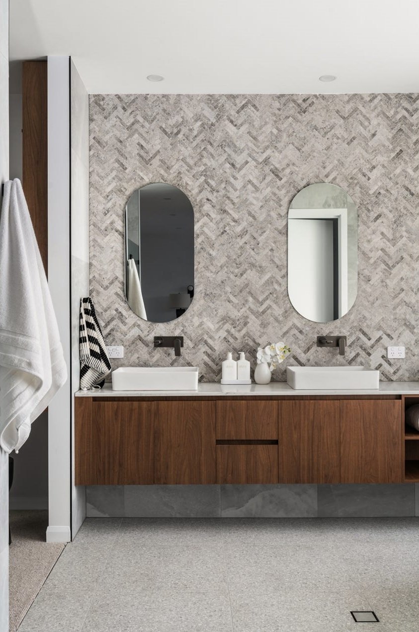Modern bathroom with double vanity, including two white vessel sinks, rectangular mirrors with rounded edges, and wooden cabinetry, against a patterned wall with chevron designs.