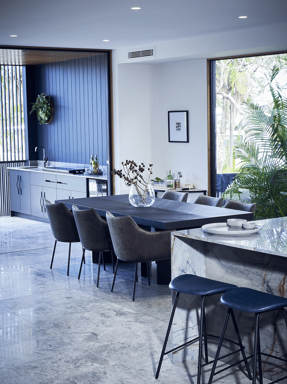 Modern kitchen and dining area with marble island, dark dining table, plush chairs, large windows, indoor plants, and minimal decor.