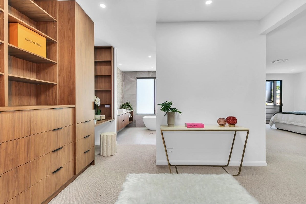 Modern interior living space with wooden shelving and a partial view of a bathroom with a bathtub, and a bedroom with a bed, all with white walls and carpeted floors.