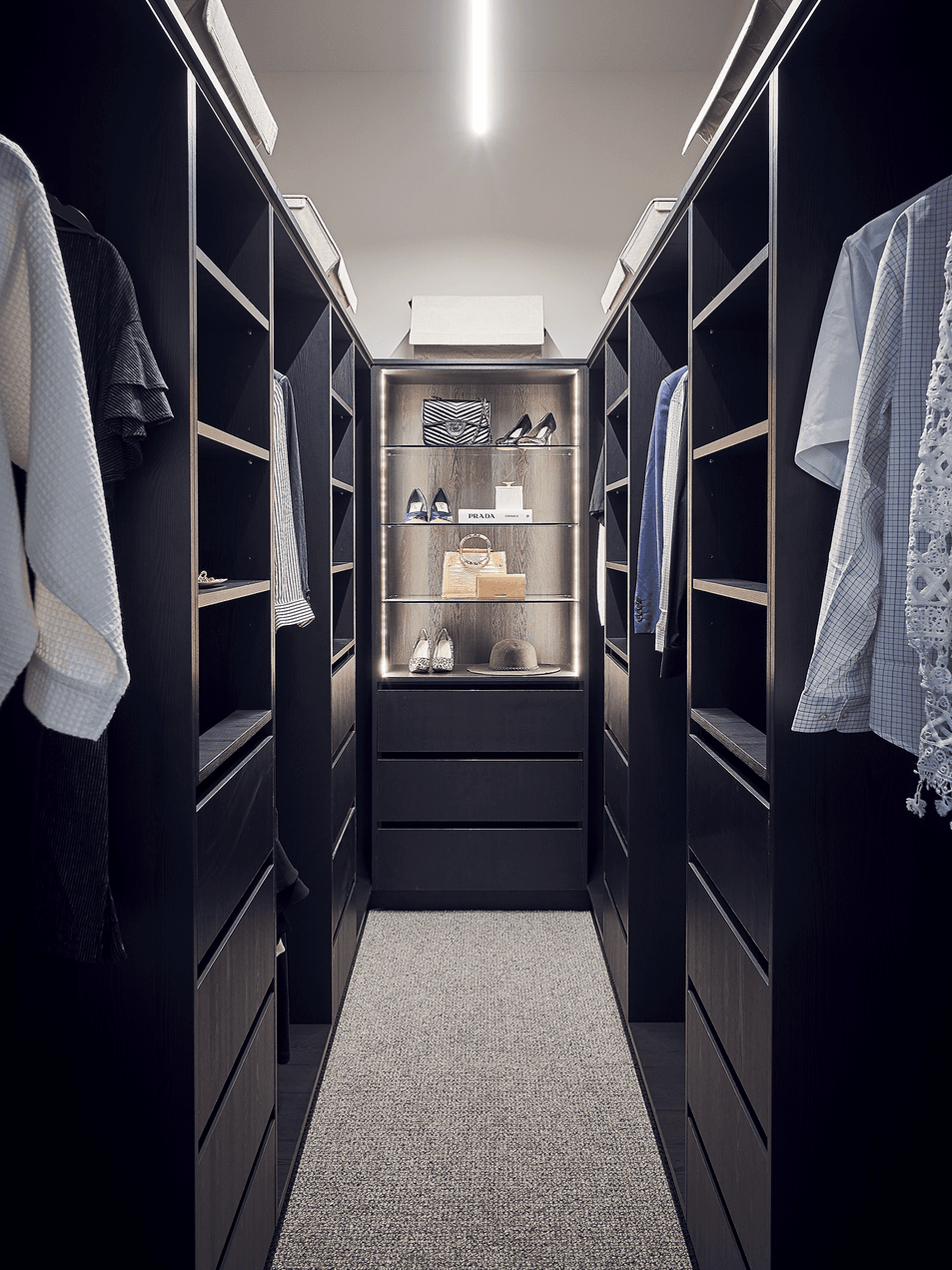 A walk-in closet with dark wooden shelving and hanging clothes, illuminated shelves with shoes, handbags, and accessories, and a carpeted floor.