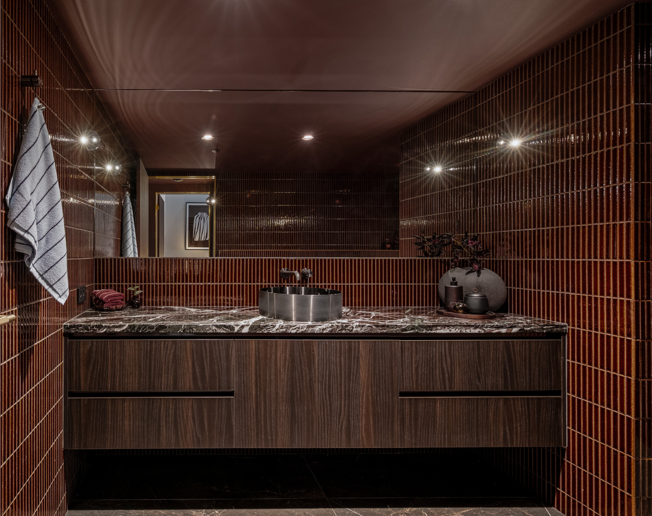 A modern bathroom with dark brown tiled walls, a marble countertop, and a round stainless steel sink. There are towels on hooks, decorative items, and a large mirror reflecting a framed artwork.