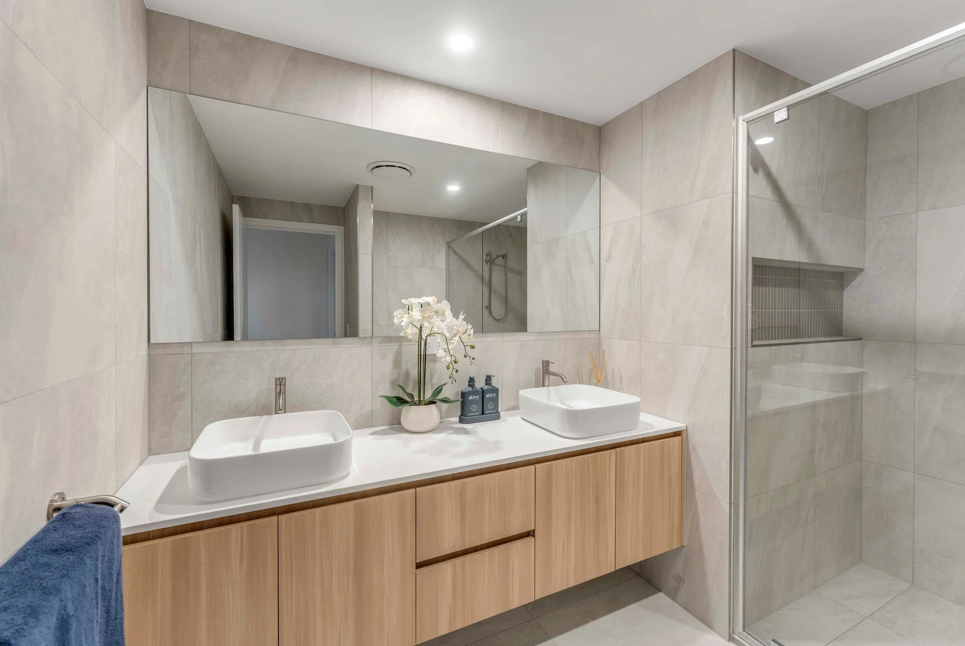 Modern bathroom with double vessel sinks, a large mirror, a wooden vanity, a potted orchid, two soap dispensers, and a walk-in shower with a glass door.