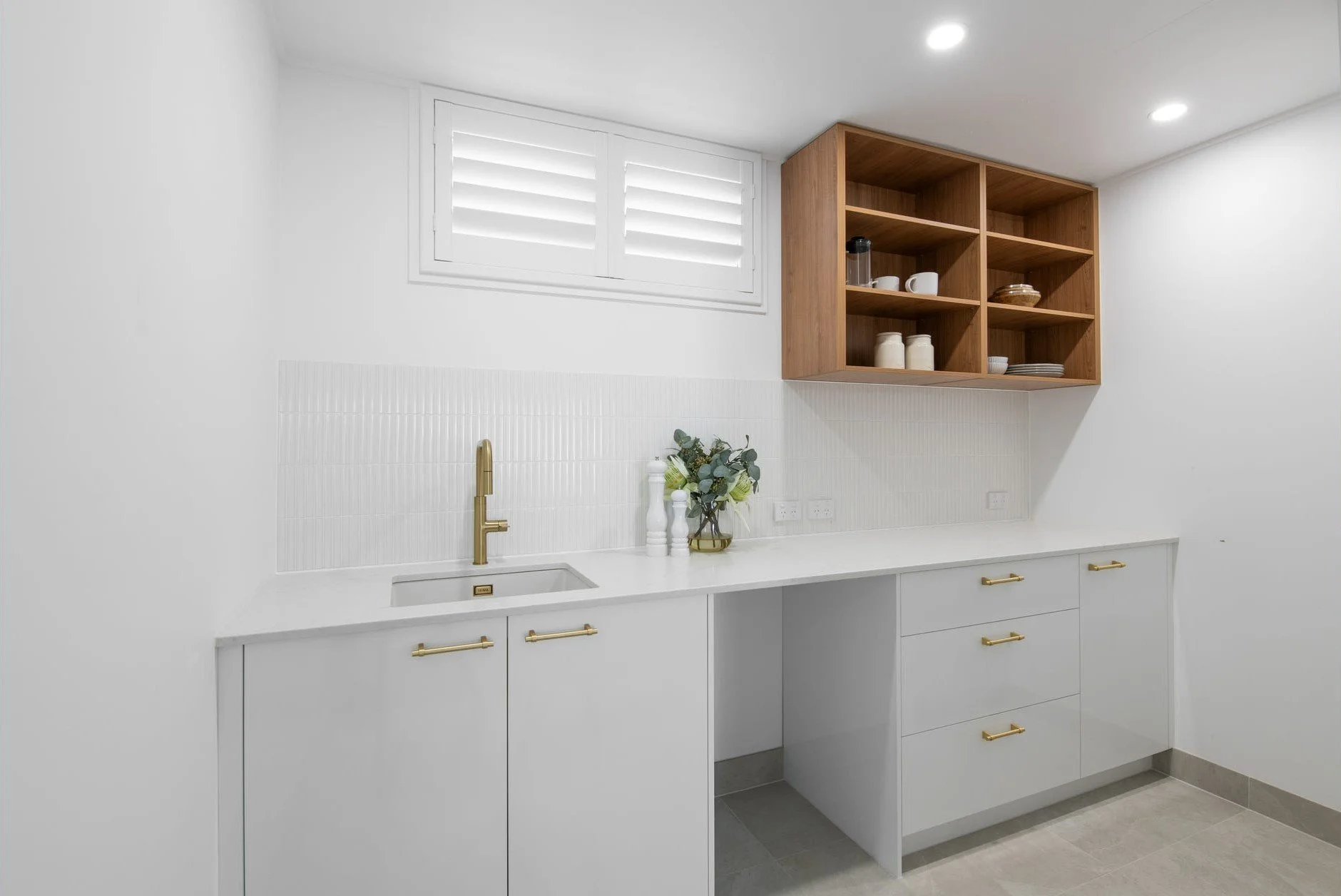 Minimalist kitchen with white cabinets, brass handles, a gold faucet, a countertop with a small sink, a vase with green foliage, an open wooden shelf with dishes, and a window with white shutters.