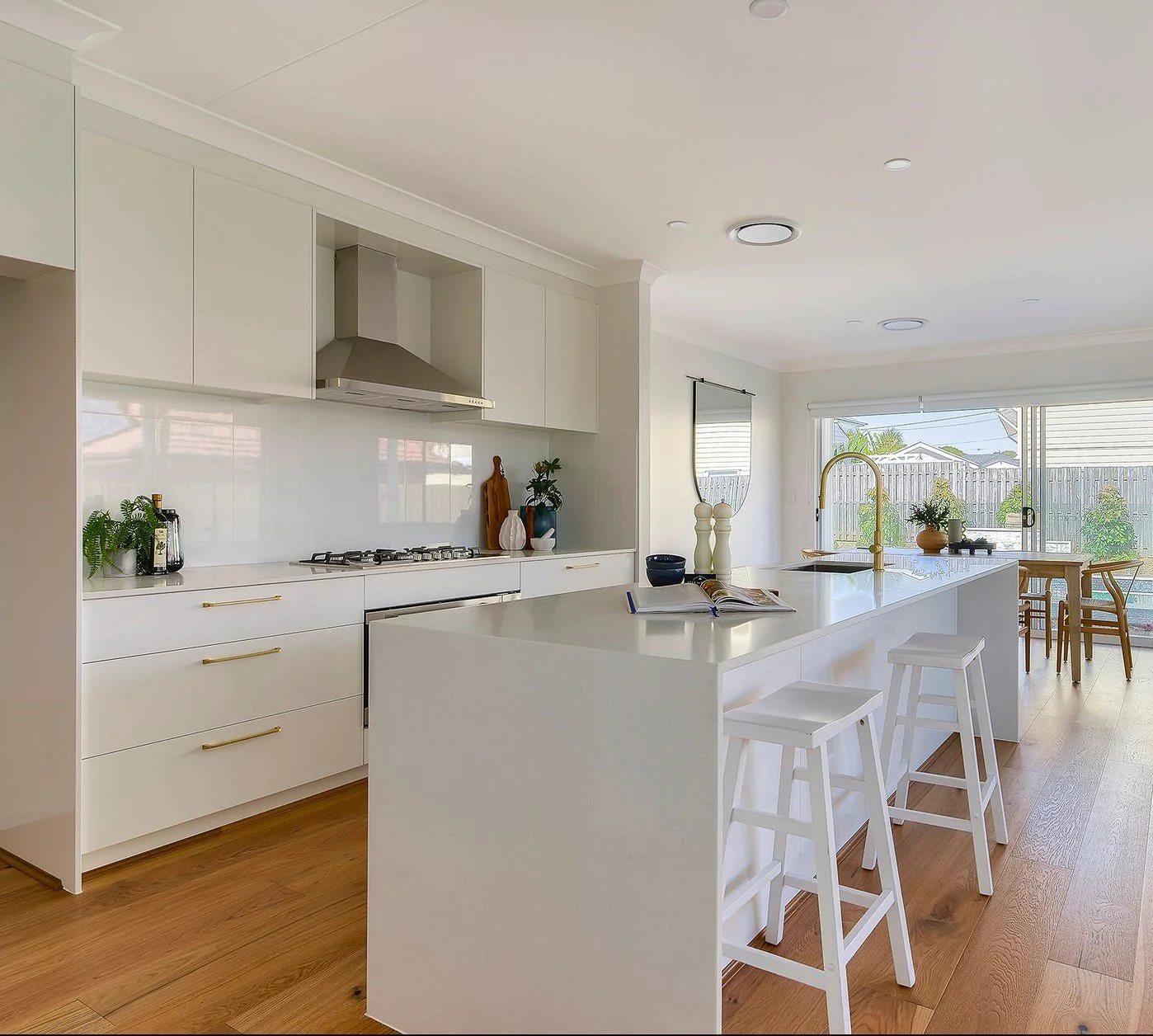 Modern white kitchen with island, bar stools, dining area, large window, and hardwood floors.