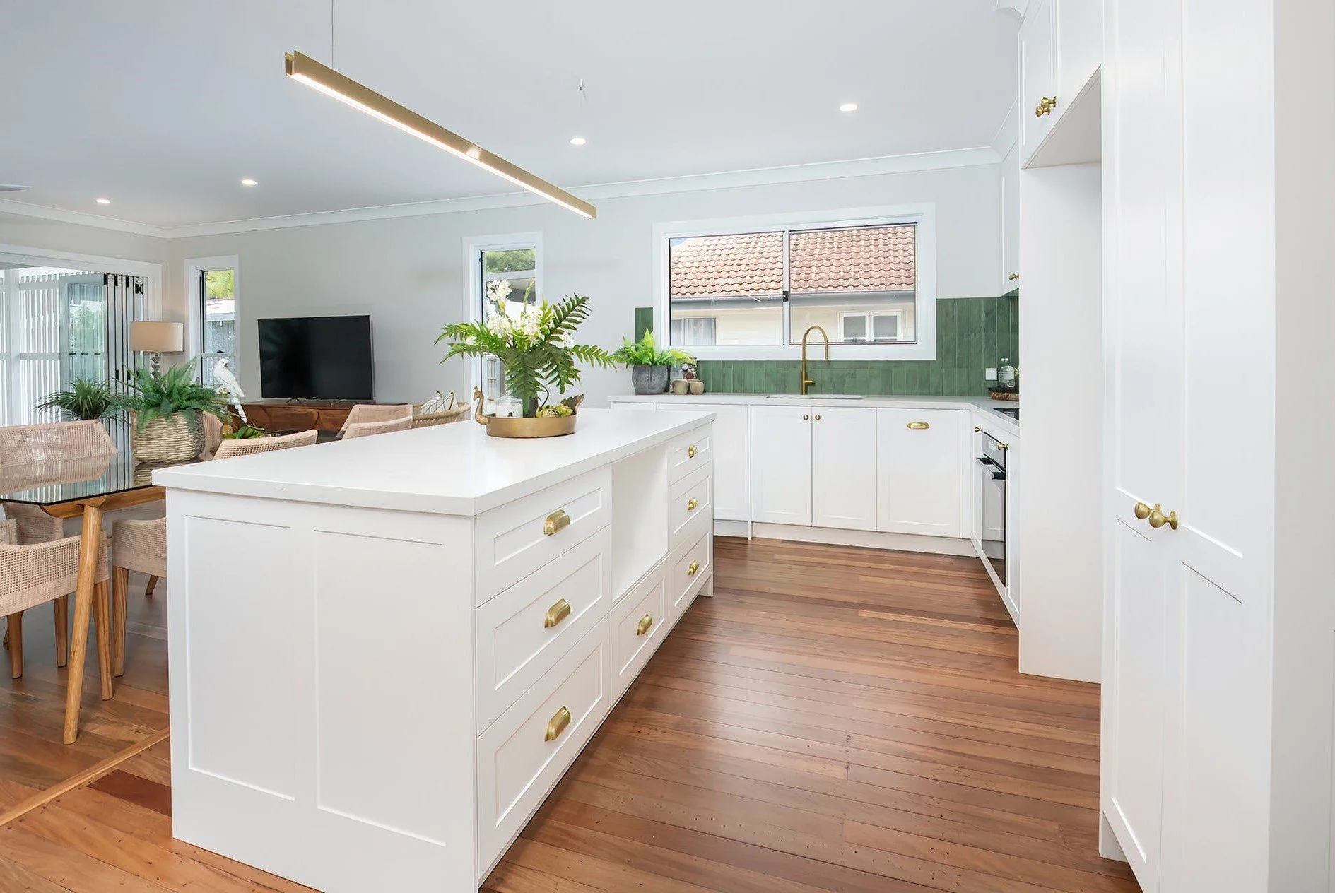 Bright, modern kitchen with white cabinets, green backsplash, wooden floors, and gold hardware, open to a dining area and living room.
