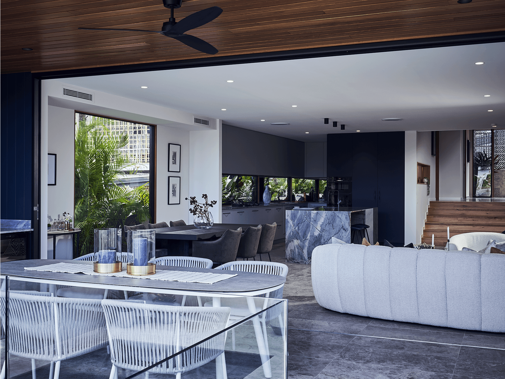 A modern open-concept living space with a dining area, a kitchen with dark cabinetry and a marble island, and a staircase with wooden steps. Large windows show greenery outside. The ceiling has recessed lighting and a black ceiling fan.