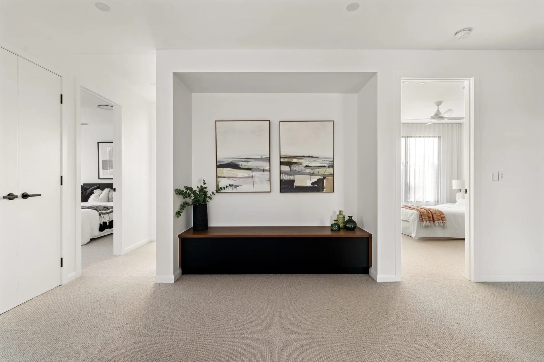 Modern living room with white walls, beige carpet, two bedrooms visible through doorways, abstract landscape paintings above a wooden console table decorated with green vases and plants.
