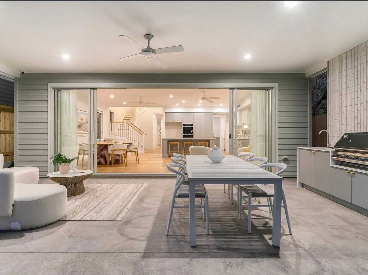 Outdoor patio with a dining table and chairs, a barbecue grill, and views into a bright indoor living and kitchen area with ceiling fans.