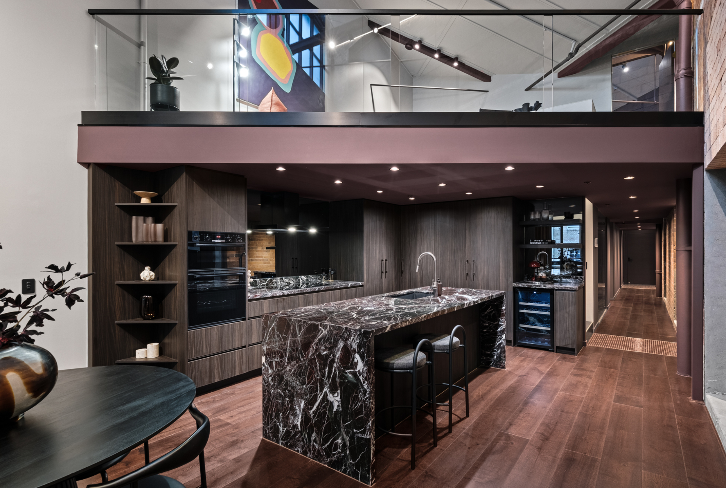 Modern kitchen with dark wood cabinets, marble island, and open shelving, featuring a hallway and upper loft with a glass railing and minimal decor.