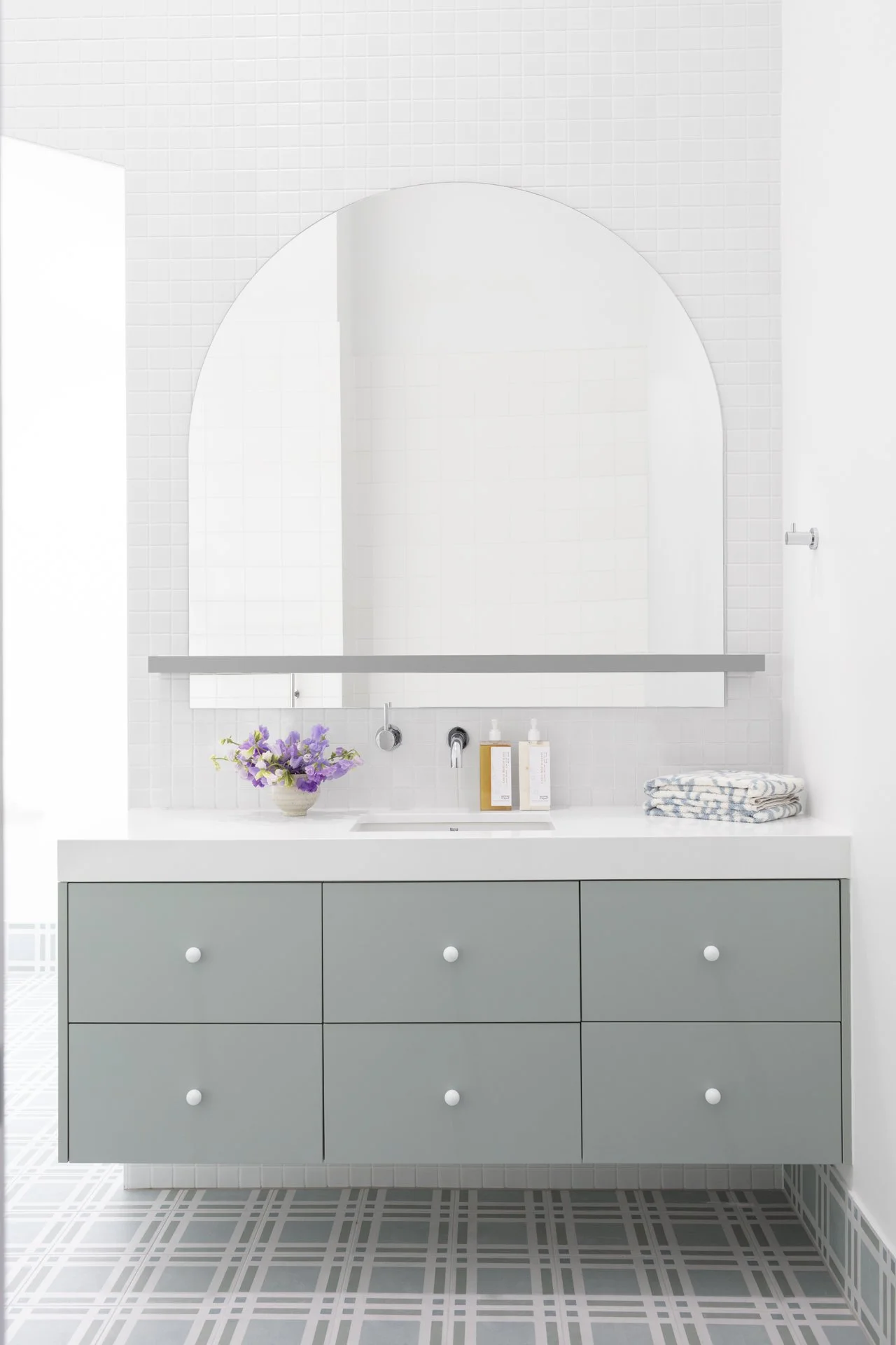 Modern bathroom with a large mirror, a lavender flower arrangement, soap and lotion bottles, and folded towels on a white countertop vanity with six drawers, white tiled walls, and patterned floor tiles.