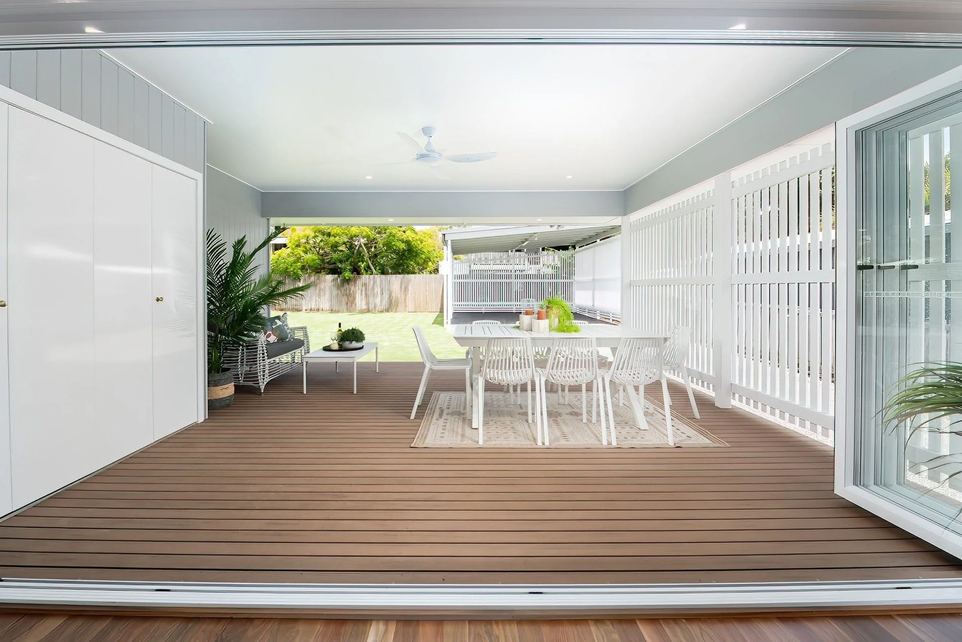 Covered outdoor patio with a wooden floor, dining table with white chairs, large sliding glass door, and a view of a grassy backyard