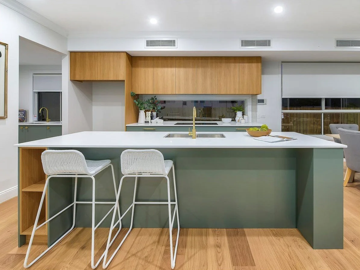 Modern kitchen with an island counter, two white bar stools, wooden cabinets, and green lower cabinets, with a window behind the sink and a bowl of green apples on the counter.