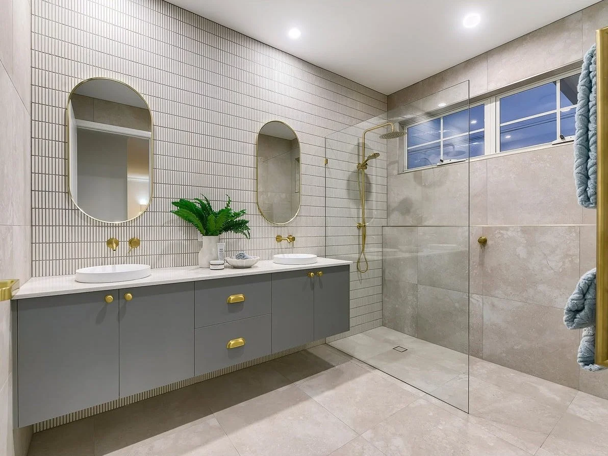 Modern bathroom featuring a double vanity with gray cabinets, gold hardware, two oval mirrors, a potted plant, and a walk-in shower with a gold showerhead and a glass partition.