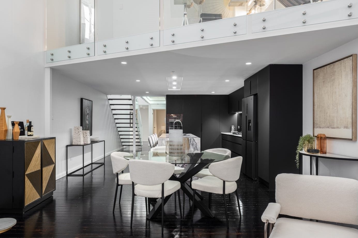 Modern open-concept living and dining area with black, white, and wood decor, black wooden flooring, a dining table with white chairs, a black kitchen with built-in appliances, and decorative items on furniture.