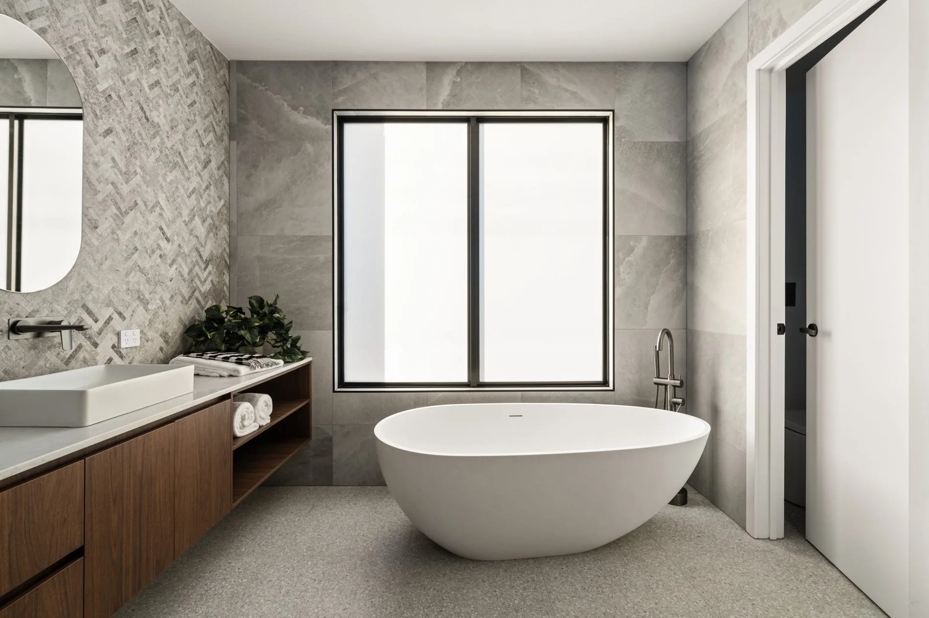 Modern bathroom with a freestanding white bathtub, large window, wooden vanity with a sink, mirror, green plant, and a partially open sliding door.