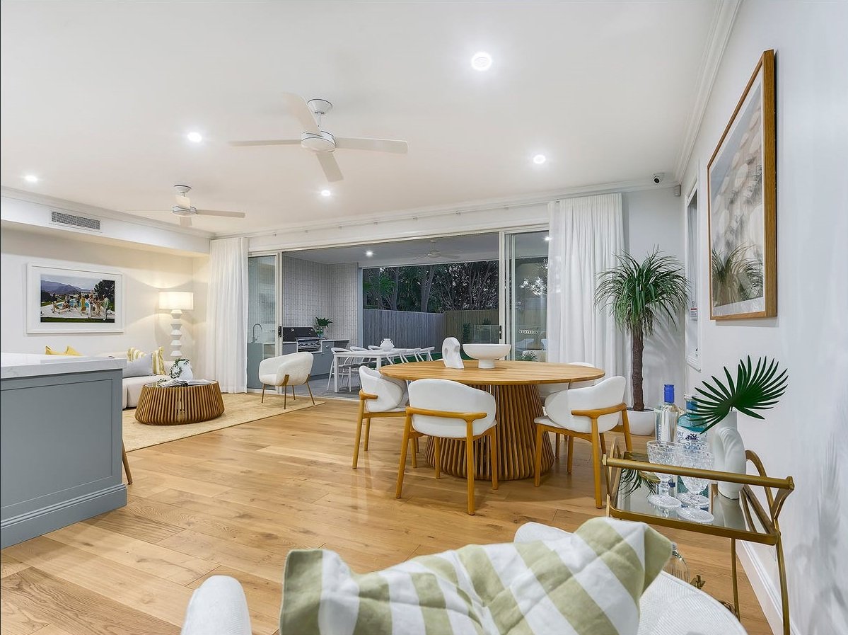 Open concept living and dining area with hardwood floors, white walls, and sliding glass doors leading to an outdoor patio. The room features a round dining table with white chairs, a white sofa with yellow accent pillows, a coffee table, houseplants