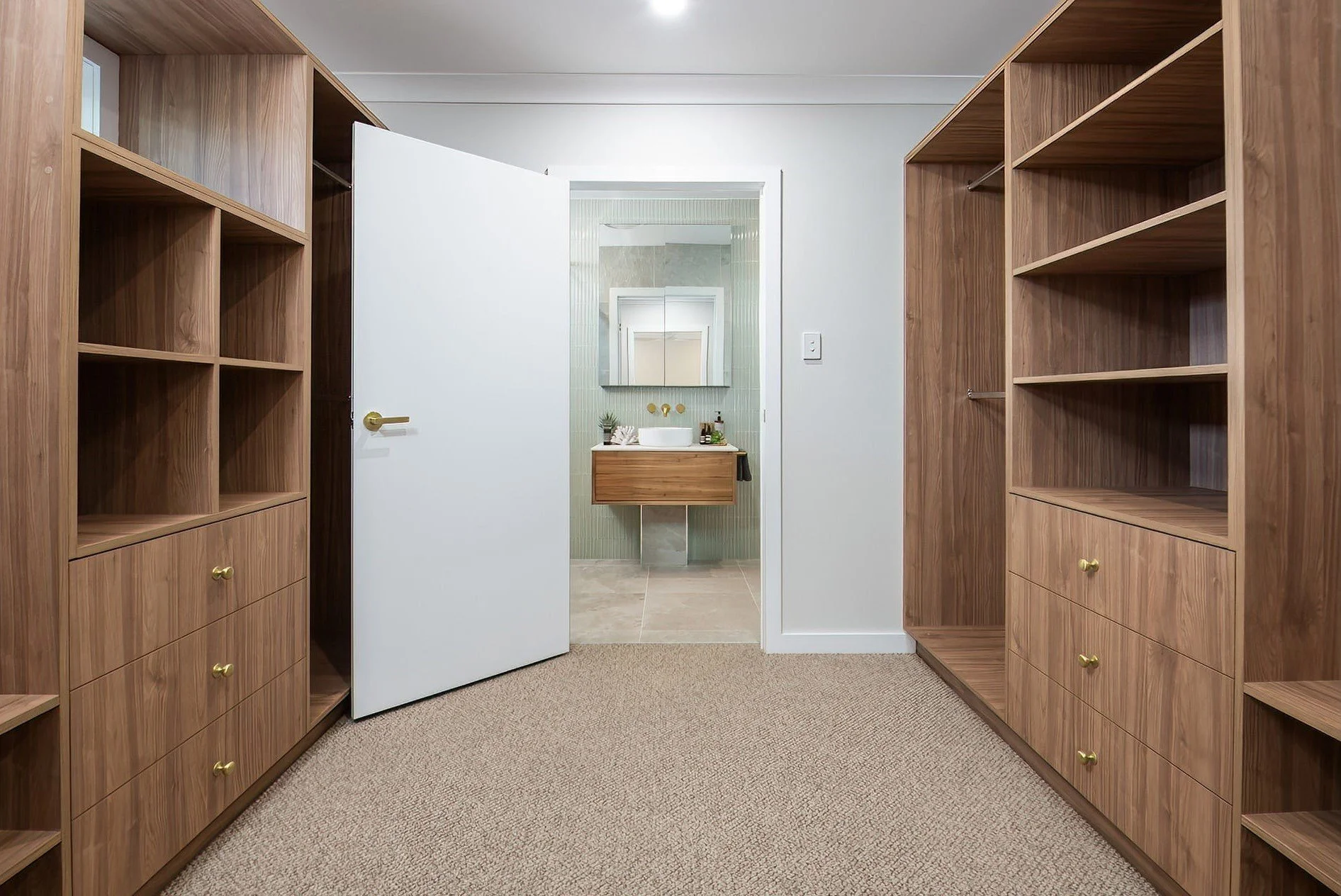 Walk-in closet with wooden shelves and drawers on both sides, open white door leading to bathroom with green tiles, sink with wooden cabinet, mirror, and small plants.