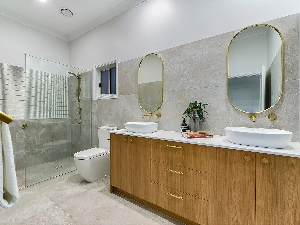 Modern bathroom with double vanity with two vessel sinks, oval mirrors with gold frames, a glass-enclosed shower with beige tiles, a toilet, a small window on the wall, and a potted plant on the countertop.