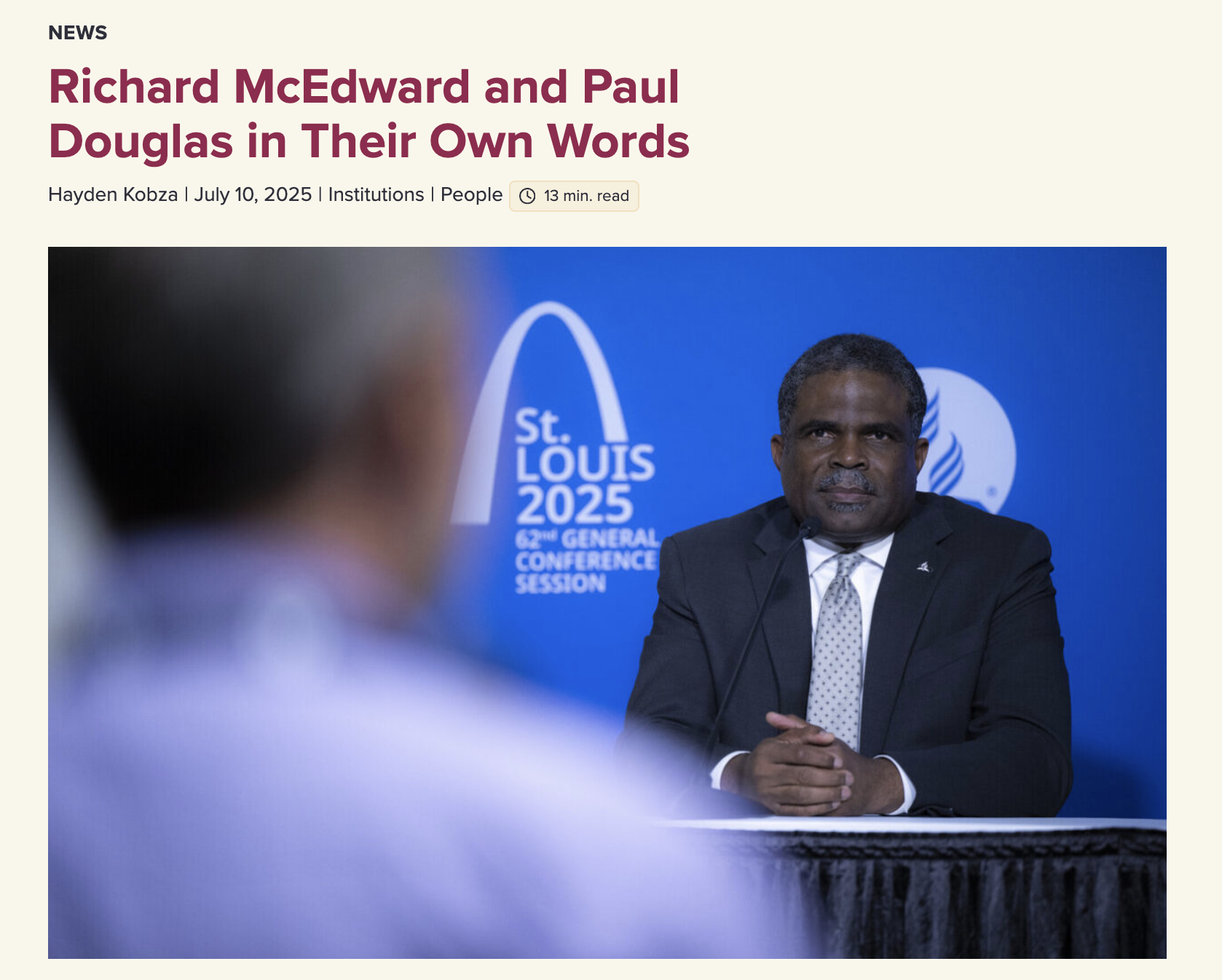 A man in a suit sitting at a table with a blue background behind him that features the St. Louis 2025 logo and text about the 62nd general conference session, with another person's blurred head in the foreground.