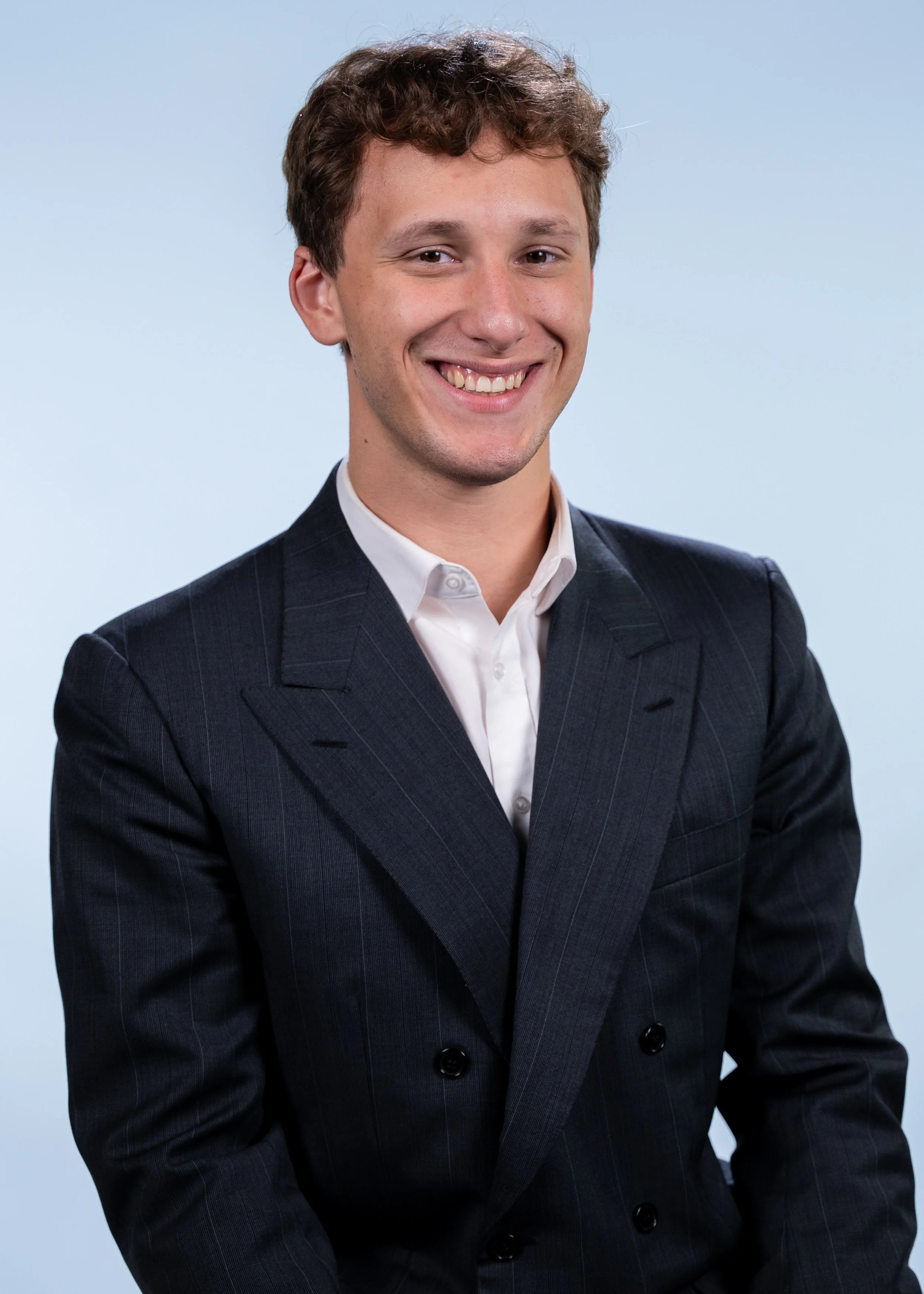 Young man with brown curly hair smiling, wearing a dark pinstripe business suit and white dress shirt, standing against a light blue background.