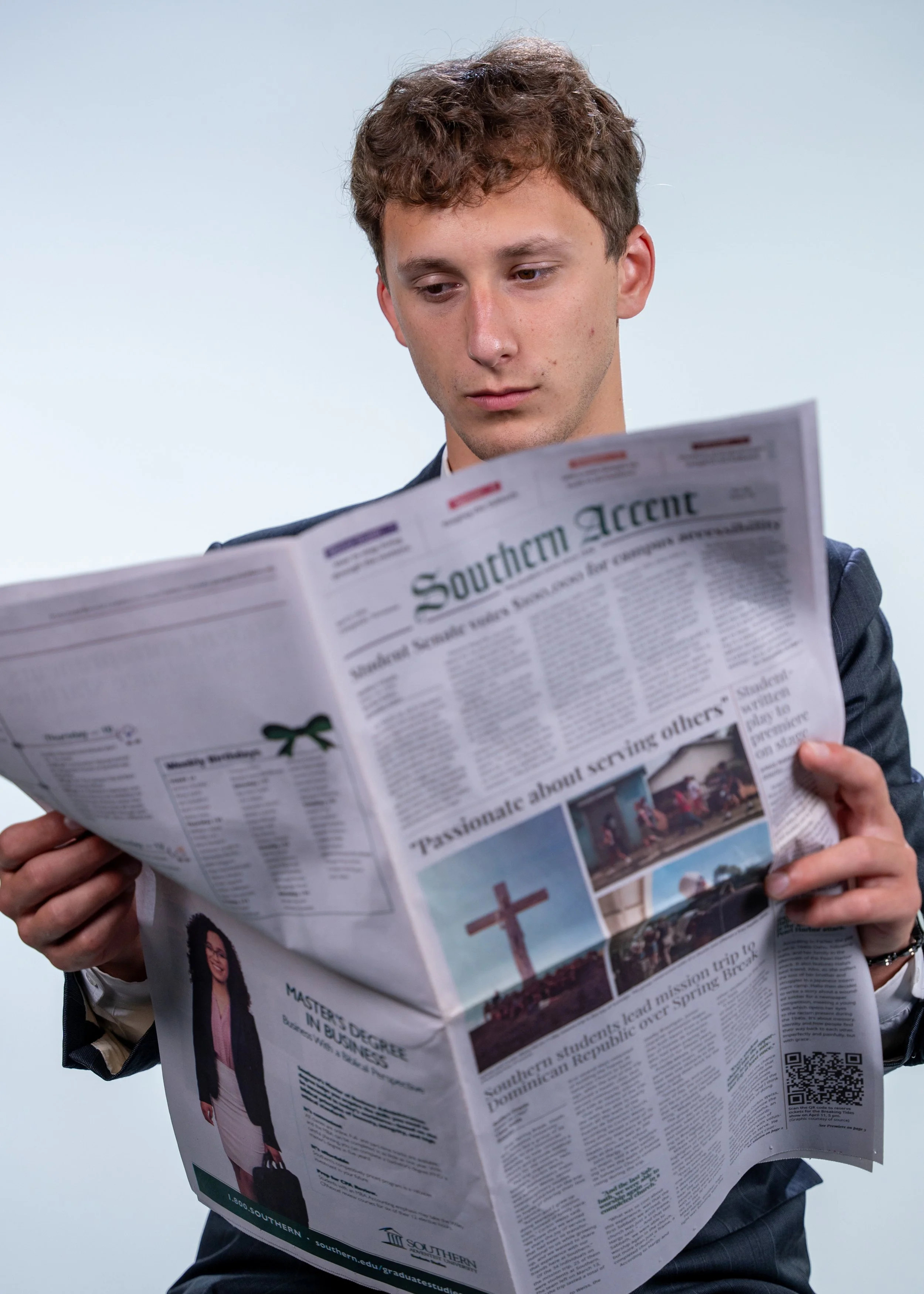 A young man in a suit reading a newspaper titled 'Southern Accent' against a plain light background.