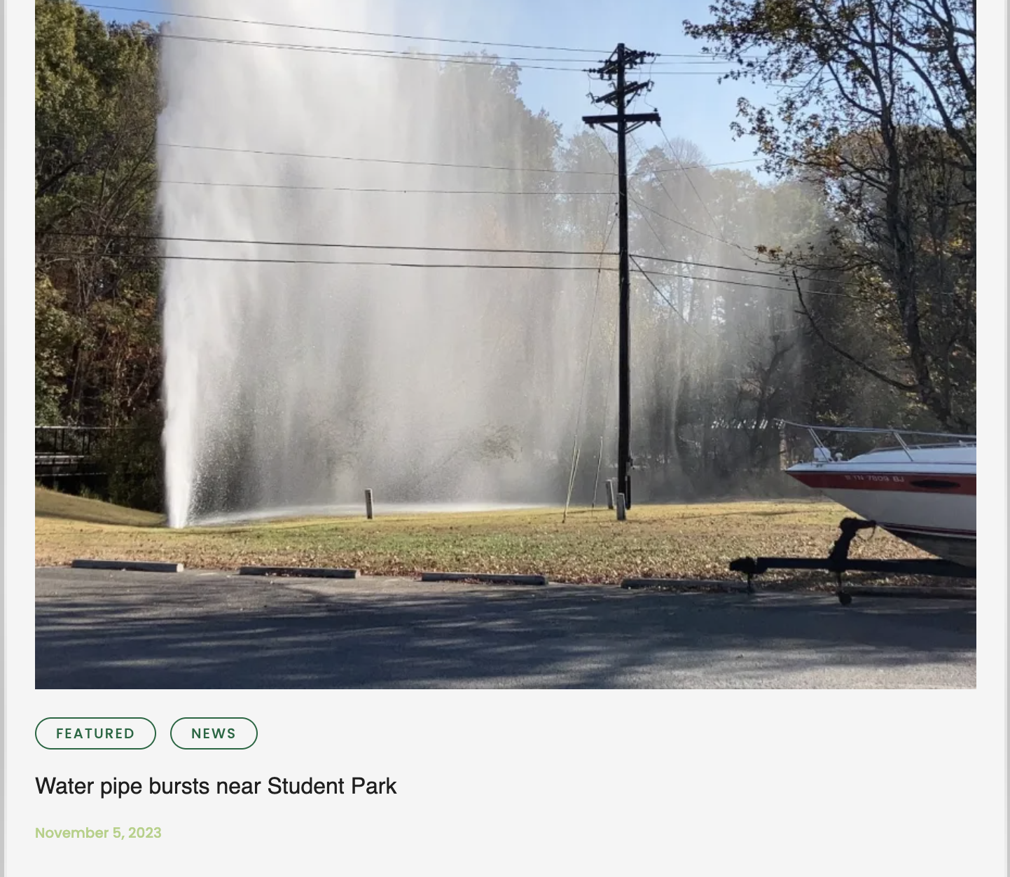 A large water pipe burst spraying water high into the air near a park, with a boat on a trailer and a power line in the foreground.
