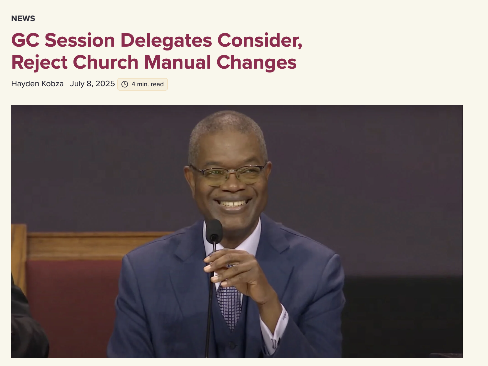 A man in a blue suit and glasses smiling while holding a microphone during a formal event or hearing.