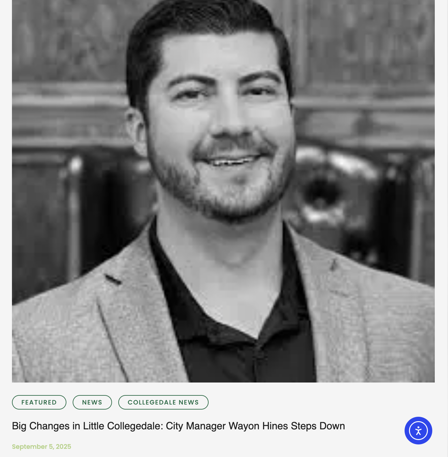 Black and white photograph of a man smiling, dressed in a light blazer and a dark shirt, sitting in a formal setting with wooden furniture in the background.