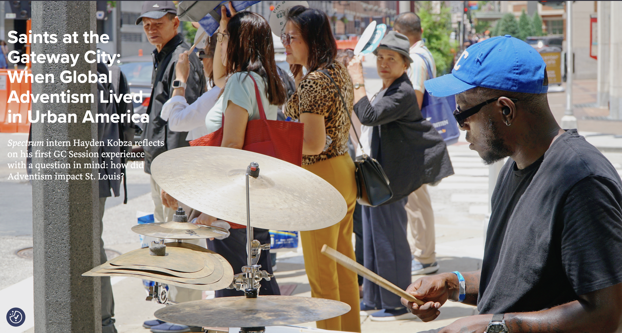 A man playing drums on a street corner with a group of people lining up in the background.