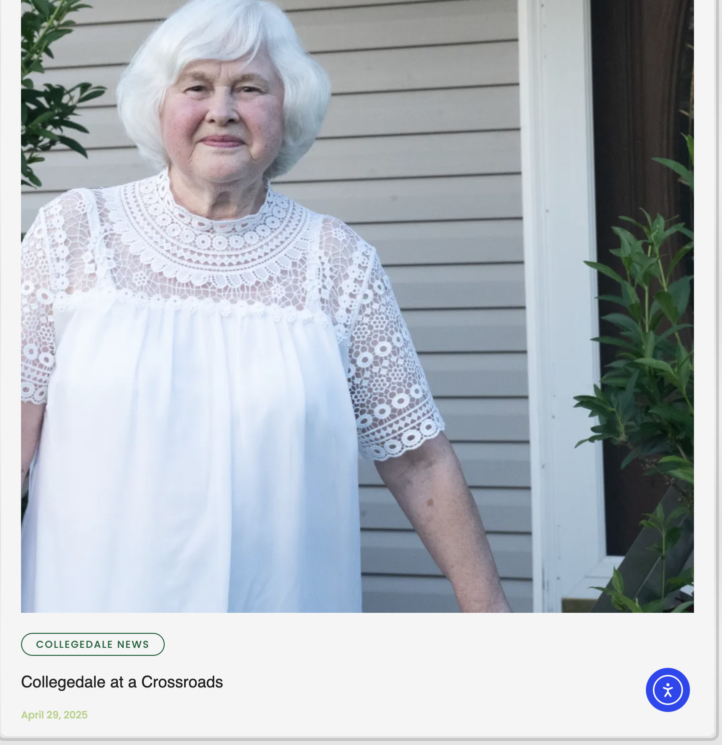 An elderly woman with white hair standing outdoors near a house with beige siding, wearing a white lacy top with detailed embroidery and a relaxed expression.