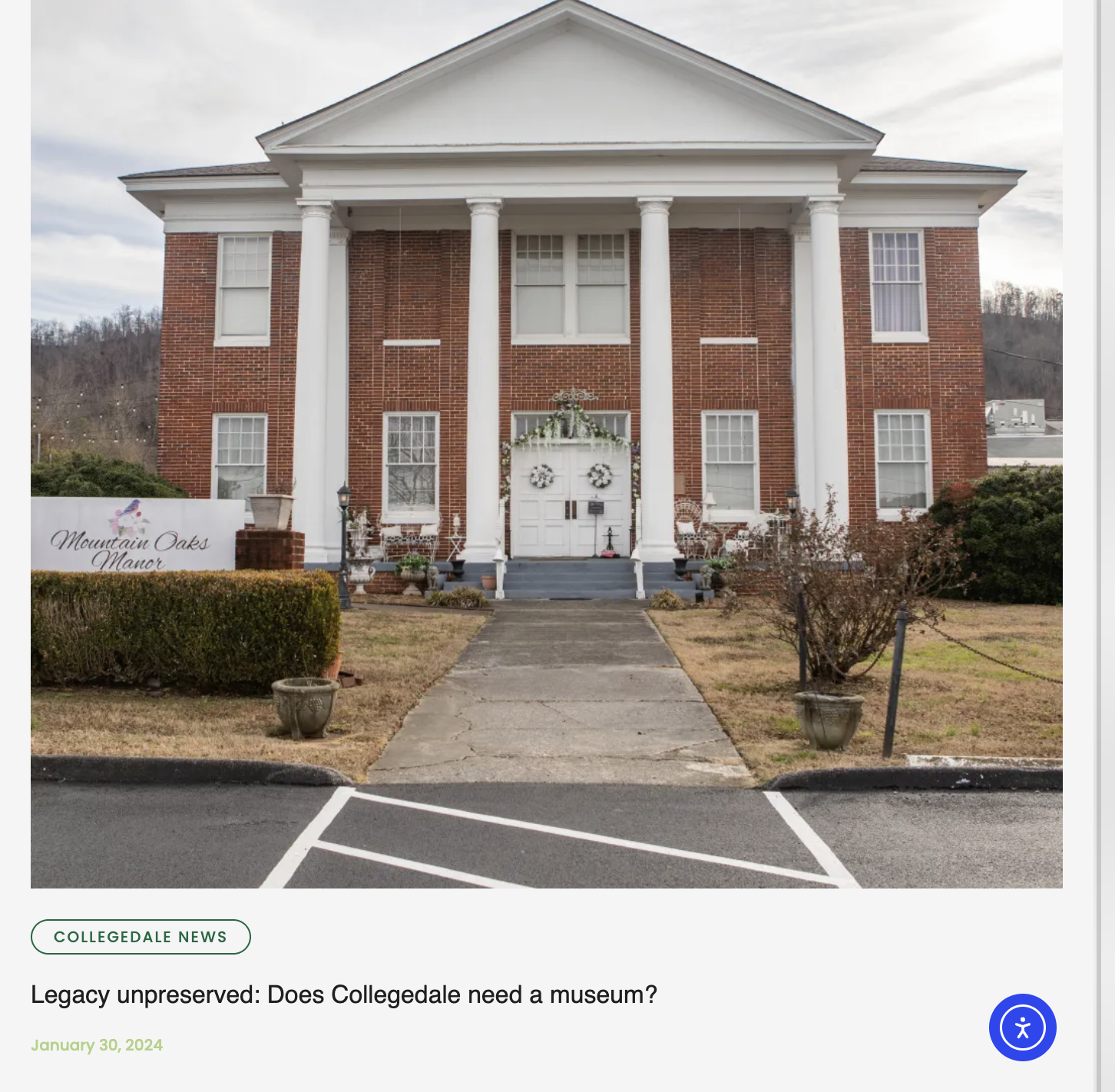 A large two-story brick building with white columns and a triangular pediment. There is a walkway leading to the front door, which is decorated with holiday wreaths and garland. Small potted plants and outdoor furniture are on the porch. A sign on th